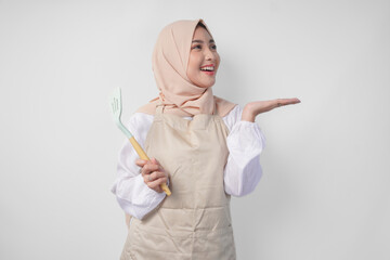 Happy young Asian Muslim woman in a veil hijab and cream apron holding spatula while pointing to the copy space beside her