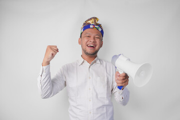 Energetic Balinese man in white shirt and traditional headdress shouting at megaphone while raising clenched fist, isolated by white background