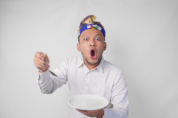 Shocked young Balinese man in white shirt and traditional headdress holding an empty plate with...