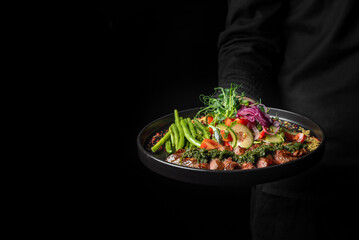 A person holds a plate of colorful, freshly prepared salad with grilled meat against a dark...