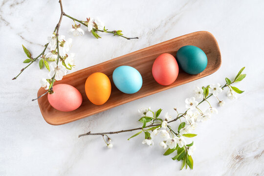 colorful chicken eggs lie in a clay plate on a light marble background surrounded by flowering branches of spring trees. easter postcard, colorful eggs, Easter composition