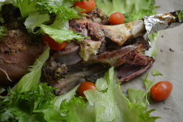 Eating, cooking delicious meat, sheep leg on the bone marinated and baked in the oven. A large piece of lamb arranged on brown parchment paper with green lettuce leaves and small cherry tomatoes.
