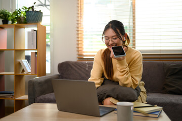 Shot of smiling young woman having phone conversation and using laptop on sofa