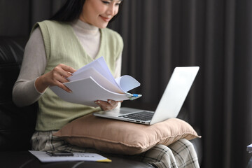 Smiling young woman sitting on couch with laptop and reading financial reports
