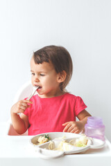 cute 1.5 year old girl sits in a high chair on a white background and learns to eat with a fork, concept of childhood and baby food. vertical photo