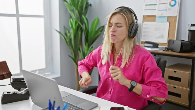 A young caucasian woman enjoys music through headphones while working at her office desktop, displaying a lively and positive work environment.