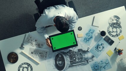 Top view of skilled engineer writing in paper while looking at tablet display green screen with turbine engine sketch design and metal gear and architectural equipment scatter around. Alimentation.