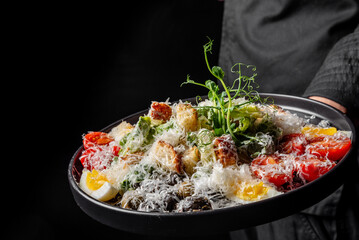A person holds a vibrant plate of fresh salad, featuring colorful vegetables, cheese, and eggs, illuminated dramatically