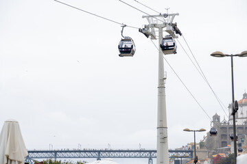 Two cable car gondolas suspended on hanging steel cables with the mechanism transporting tourists with the Don Luis I Bridge in the background, under a cloudy white sky in Porto. - obrazy, fototapety, plakaty