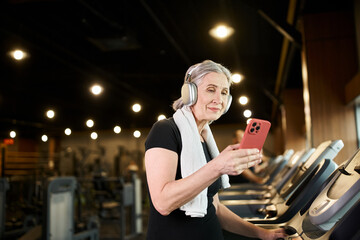 jolly mature woman with towel on shoulders and headphones holding phone and exercising on treadmill