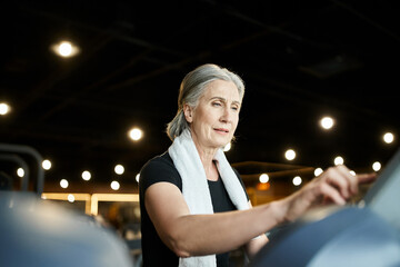athletic jolly senior woman in sporty attire with towel on shoulders running on treadmill in gym