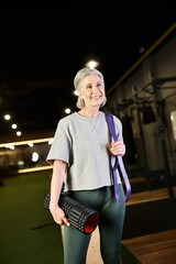 cheerful appealing mature woman posing in gym with fitness expander and weight bag and looking away