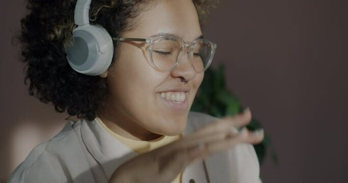 Close-up of joyful African American woman wearing wireless headphones listening to music and dancing relaxing indoors at home. Youth culture and entertainment concept.