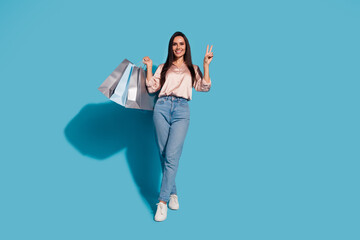 Full size photo of good mood woman dressed silk shirt jeans pants holding shopping bags show v-sign isolated on blue color background