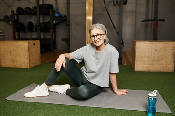 positive beautiful mature woman in sportswear with glasses sitting on floor and smiling at camera
