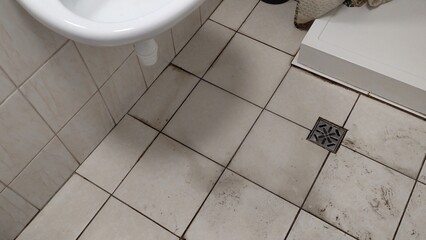 Old dirty tiled bathroom with shower and dirty floor with traces of shoes in a cheap hostel or...