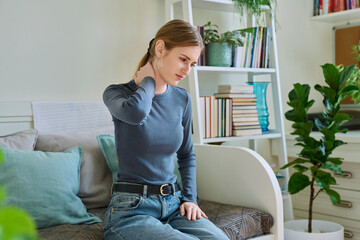 Young teenage female experiencing neck pain sitting on couch at home