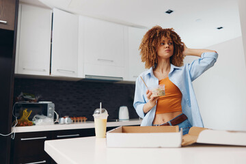 Attractive woman with curly hair standing in modern kitchen with box of food in front of her