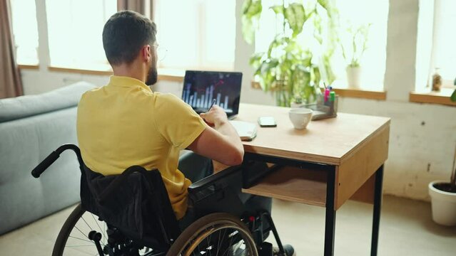 Young man sitting in wheelchair at home at desk with laptop and working with financial graph. Remote working. Financial planning, analyzing. Concept of healthcare, lifestyle, motivation, wellness