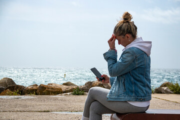 Blonde woman with bun using her mobile phone while enjoying the sea sitting on a bench on the...