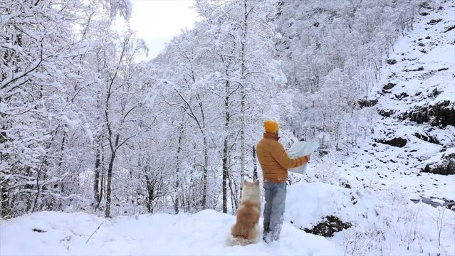 A man in a yellow beanie attentively reads a map in a snowy landscape with his loyal dog by his side. Winter Trekking: Man with Dog Reading Map in Snow