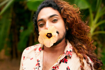 Dark-skinned man with curly long hair poses in nature, flower in mouth expressing confidence,...
