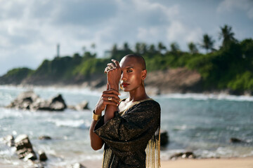 Flamboyant queer black male in luxury gown poses on scenic ocean beach. Trans sexual ethnic fashion...