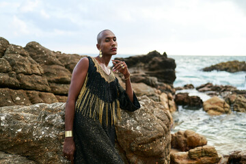 Queer black person in luxury dress sits on rocks in ocean. Lgbtq ethnic fashion model wearing...