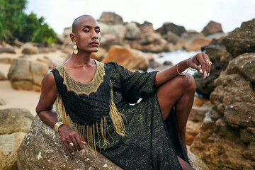 Epatage lgbtq black man poses on scenic ocean beach looks at camera demonstrates jewellery. Androgynous ethnic fashion model in posh dress, jewelry looks at camera, sits on stone. Pride month. - obrazy, fototapety, plakaty