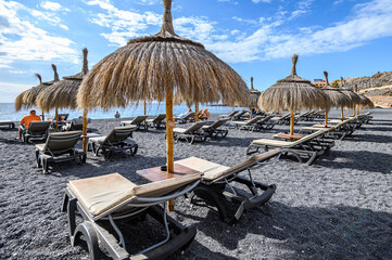 straw beach umbrella with blue sky, sunbeds on the black volcanic beach of La Caletta. Tenerife,...
