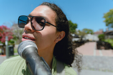 Wide angle close-up portrait of young man with long curly hair singing with microphone on street.