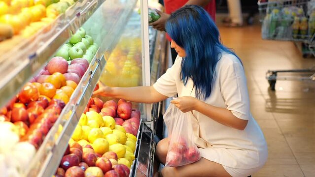 Tourist woman choosing best apples from shelf, buying imported fruits at Asian supermarket. She chooses one fruit and examines it carefully; if she is satisfied, she puts apple into plastic bag