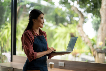 Cheerful barista in apron using a laptop, likely managing online orders or cafe operations, with natural backdrop.