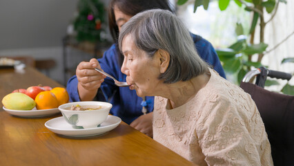 Caregiver feeding elderly woman with soup in dining room.