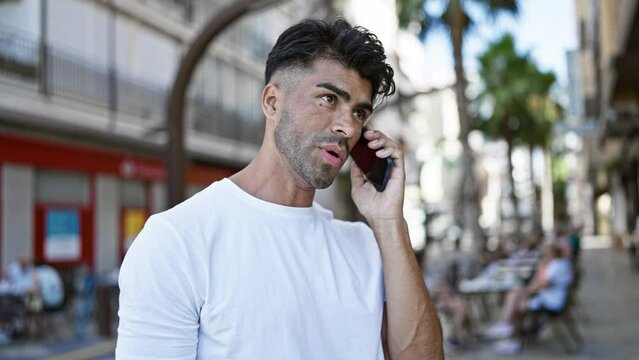 A handsome young hispanic man in casual attire attentively speaking on a smartphone while walking outdoors on a vivid city street.