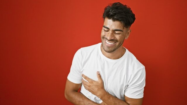 A young hispanic man with a beard smiles and points at the camera against an isolated red background