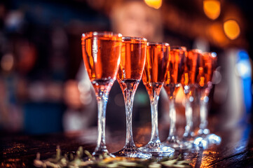 A row of six wine glasses are filled with red liquid. The glasses are arranged on a bar counter, with some of them placed closer to the edge. Concept of celebration or indulgence