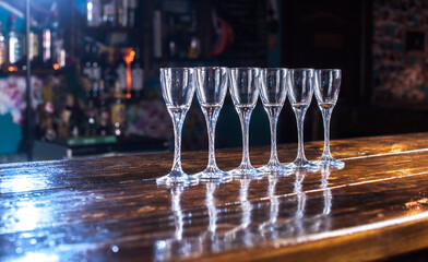 A row of six wine glasses are sitting on a wooden bar counter. The glasses are all clear and are...
