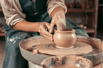 A potter is making a clay pot on a wheel. The potter is wearing a blue apron and has his hands covered in clay. Concept of focus and concentration as the potter works on his craft