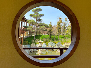 A round window in the wall overlooking a beautiful landscape. Japanese garden with a pond, stones and trees.