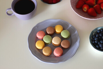Plate of pastel macarons, cookies and chocolate, cup of tea of coffee, glass of bubble water, various berries, books and accessories on the table. Selective focus, pastel colors.