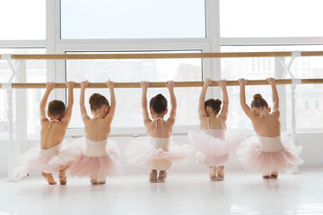 Line of little girls, dancers doing ballet steps at studio barre against window with city view....