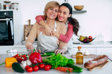 Smiling mature mother and her expressive lovely adult daughter cutting vegetables for a vegan salad together. Mid-adult woman and cute girl are preparing proper healthy meal on domestic kitchen - 777365619