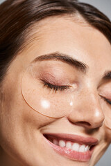 A young Caucasian woman with brunette hair applies clear under eye patches in a studio setting.