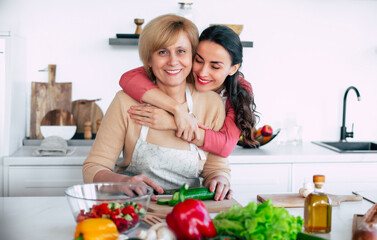 Smiling mature mother and her expressive lovely adult daughter cutting vegetables for a vegan salad together. Mid-adult woman and cute girl are preparing proper healthy meal on domestic kitchen - 777365663