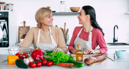Smiling mature mother and her expressive lovely adult daughter cutting vegetables for a vegan salad together. Mid-adult woman and cute girl are preparing proper healthy meal on domestic kitchen - 777365667