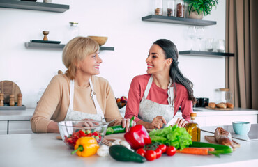 Smiling mature mother and her expressive lovely adult daughter cutting vegetables for a vegan salad together. Mid-adult woman and cute girl are preparing proper healthy meal on domestic kitchen