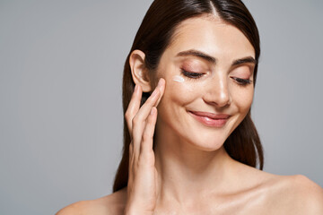A young Caucasian woman with brunette hair smiling and gently touching her face with cream