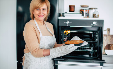 Close up image of smiling mature female in apron and kitchen gloves holds and shows her vegan cake or apple pie from oven in modern kitchen on the camera.