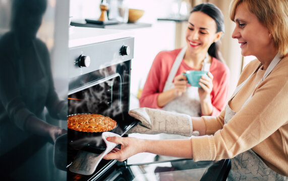 Happy lovely daughter with her mature mom baking and using kitchen gloves for to take apple vegan pie out of the oven in kitchen.